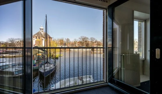 Uitzicht vanaf een balkon aan de Hoogte Kadijk op een gracht met aangemeerde boten en een historisch gebouw op de achtergrond.